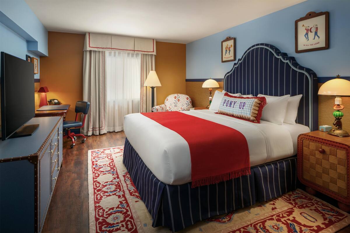 A hotel room with a large bed, two bedside tables with lamps, a desk with a chair, a television, and a window with curtains.