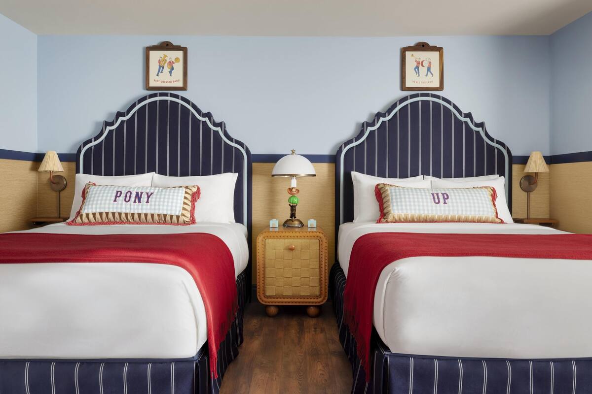 A hotel room with two beds, each with a headboard and a pillow with a name. There is a nightstand with a lamp and a small table with a decorative item between the beds.