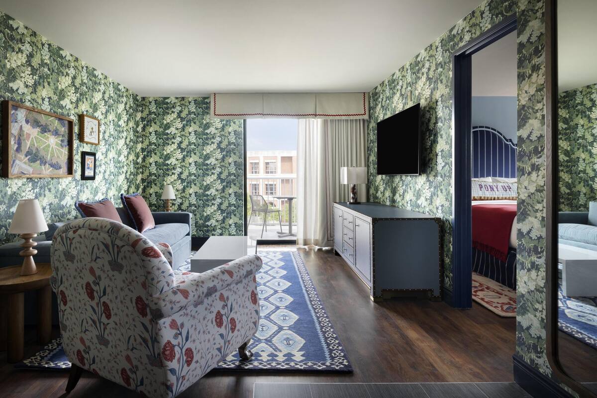 A hotel room with floral wallpaper, a sofa, a chair, a TV, a desk, and a view of the outside.