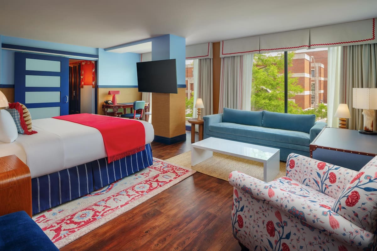 A hotel room with a large bed, a blue sofa, a floral armchair, a TV, and a view of a building and trees.