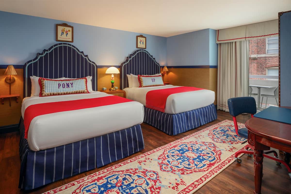 A hotel room with two beds, a dining table, and a view of a brick building.