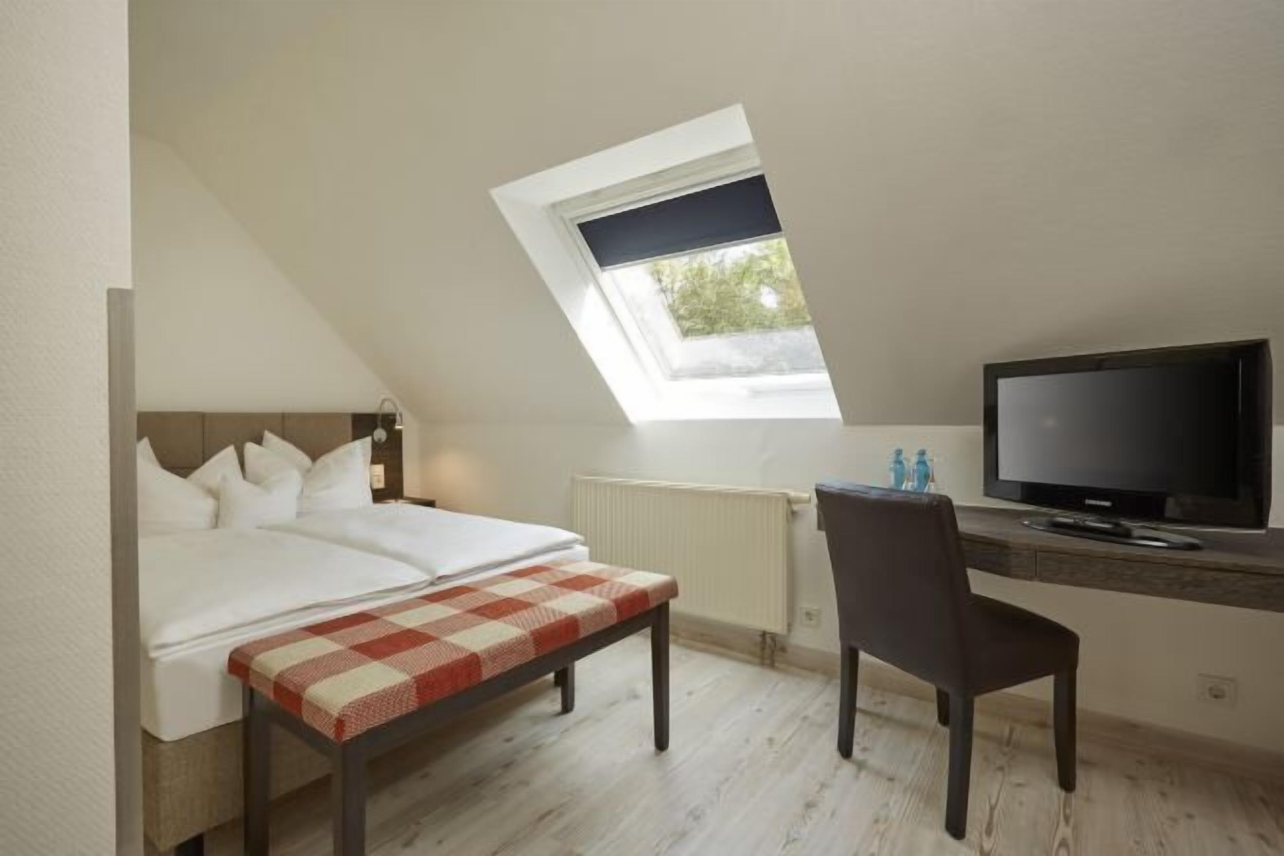 Un dormitorio con tragaluz, una cama, un banco, un escritorio con televisor y una silla.