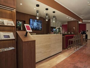 Zona de recepción de un hotel con un mostrador de madera, un televisor montado en la pared y una zona de bar con taburetes.