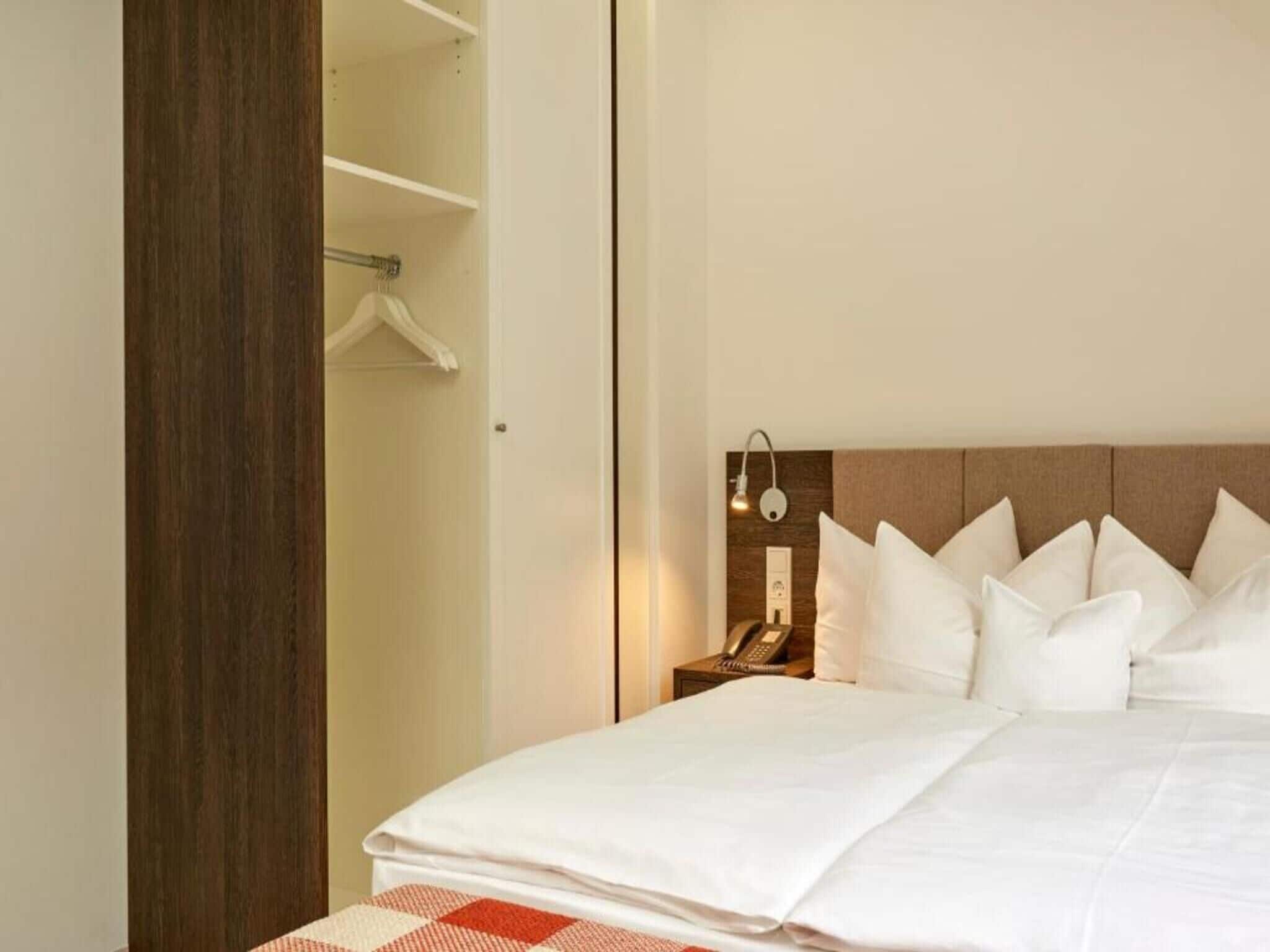 A hotel room with a bed, a bedside table with a lamp, a telephone, and a wardrobe.