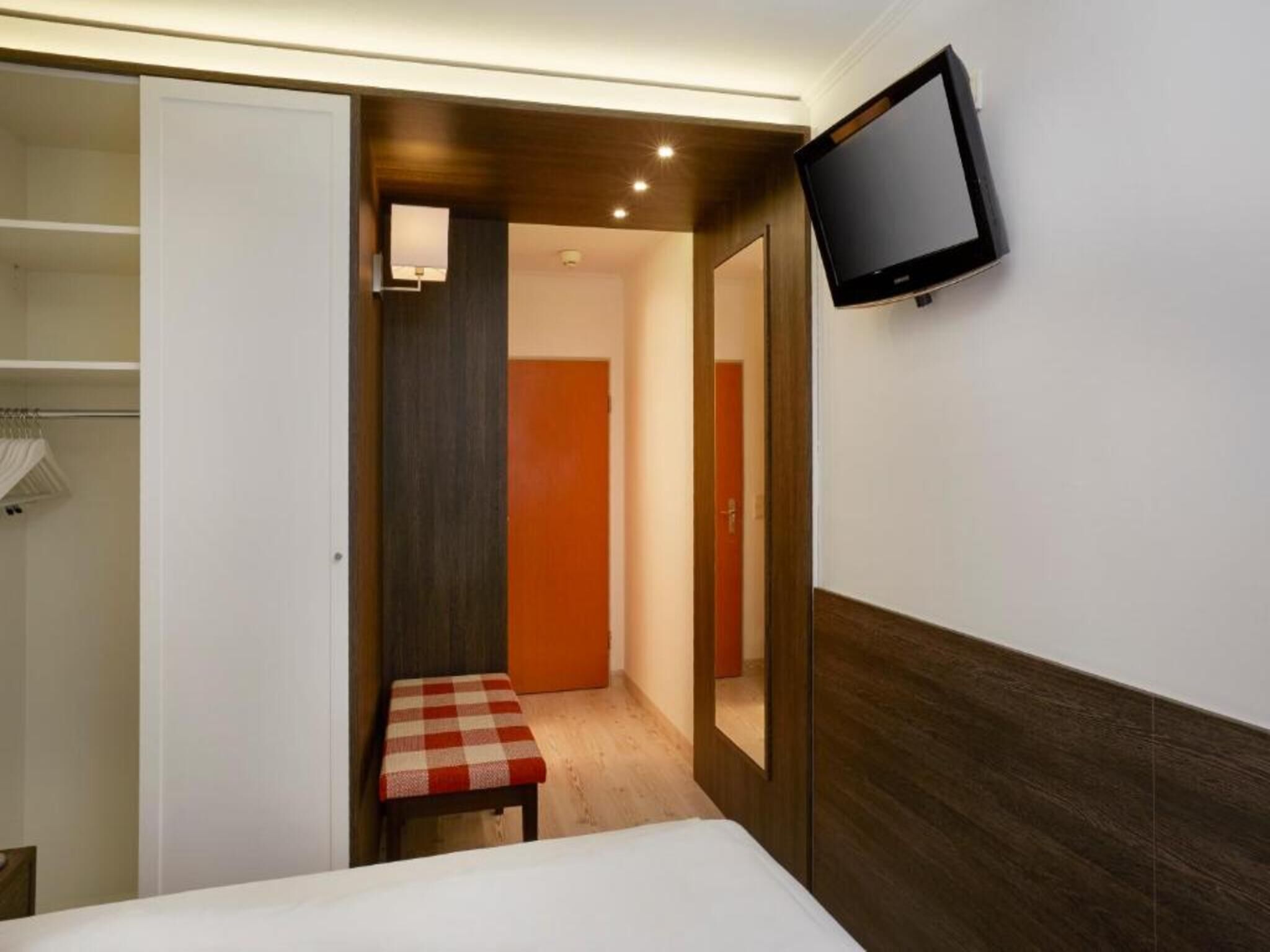 Habitación de hotel con cama, televisor montado en la pared, una mesita con mantel a cuadros y un armario con ropa colgada.