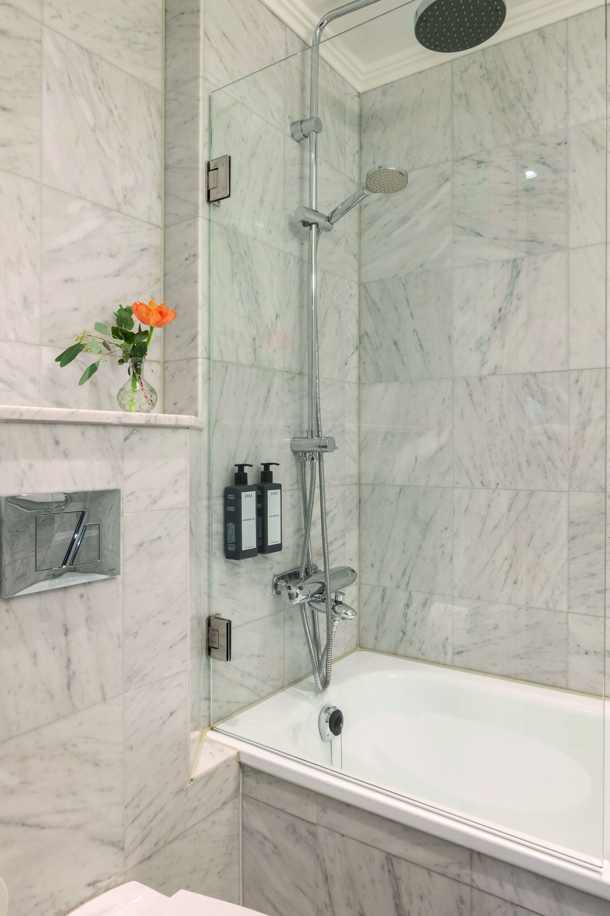 Un baño moderno con ducha envuelta en vidrio, una bañera blanca, paredes de mármol y un estante pequeño con artículos de aseo.