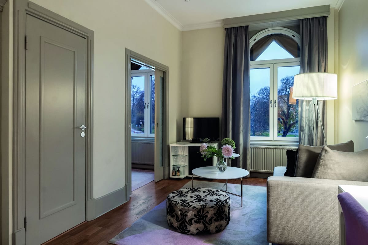 A modern living room with a sofa, ottoman, and a small table with flowers. There is a TV and a lamp on a side table.