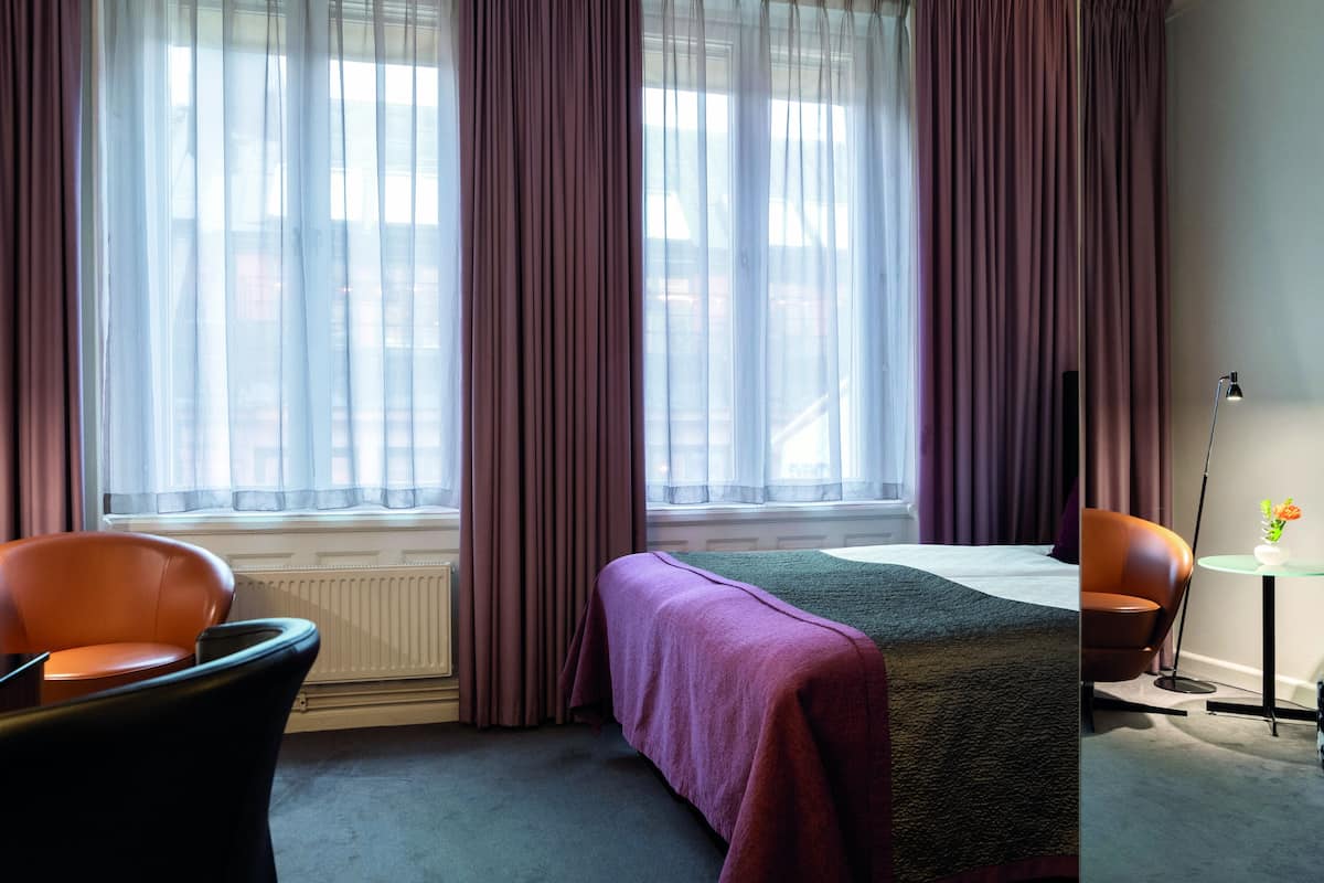 A hotel room with a bed, two chairs, a small table with a vase, and a radiator.