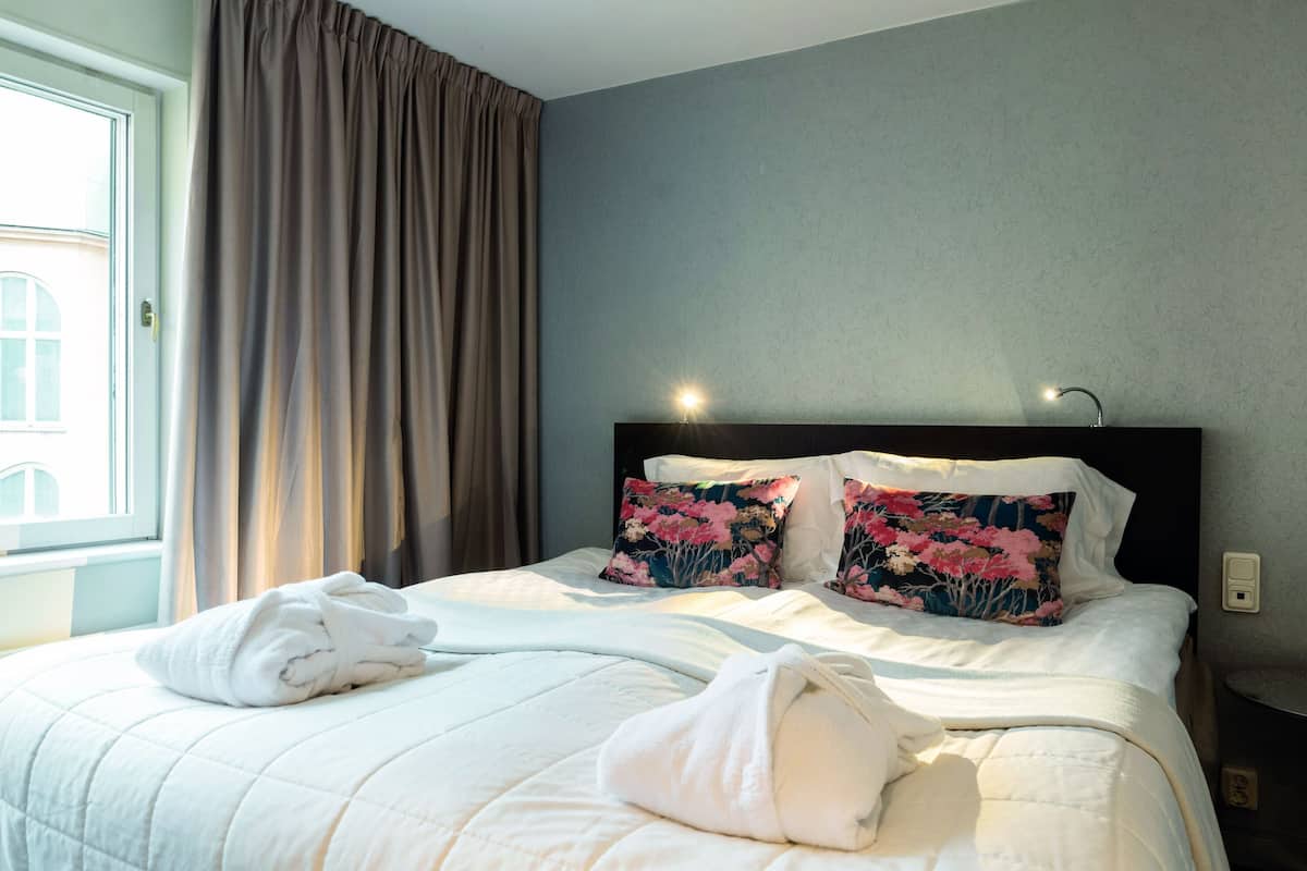 A neatly made bed with white linens and floral pillows, a window with curtains, and a wall-mounted light fixture.