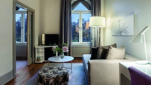 Moderni hotellihuone, jossa on sohva, pieni pöytä maljakko täynnä kukkia, televisio ja ikkuna, jossa on verhot.