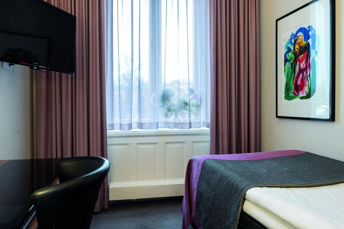 A hotel room with a bed, a desk, a chair, a window with curtains, and a painting on the wall.