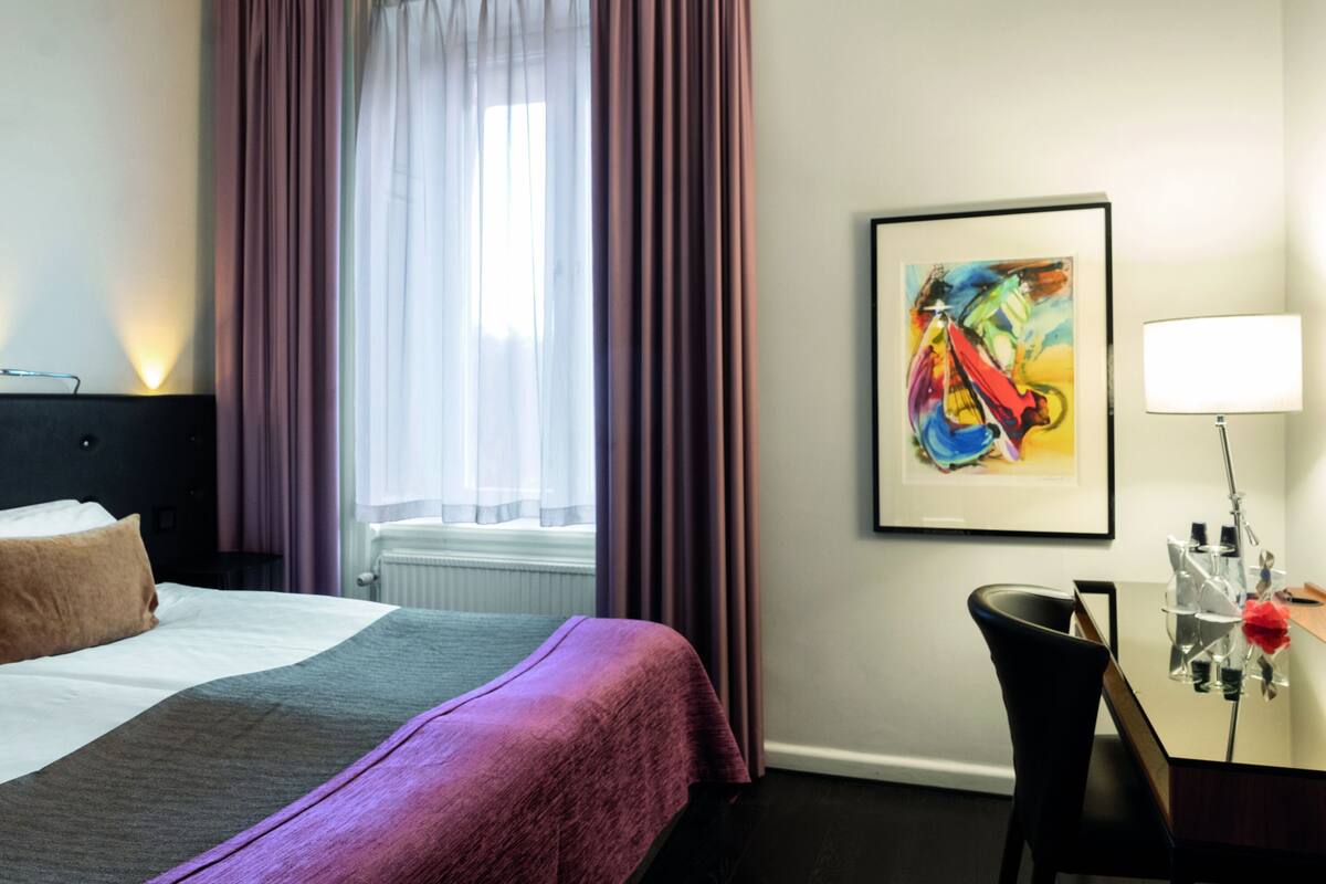 A hotel room with a bed, a desk with a chair, a window with curtains, and a framed artwork on the wall.