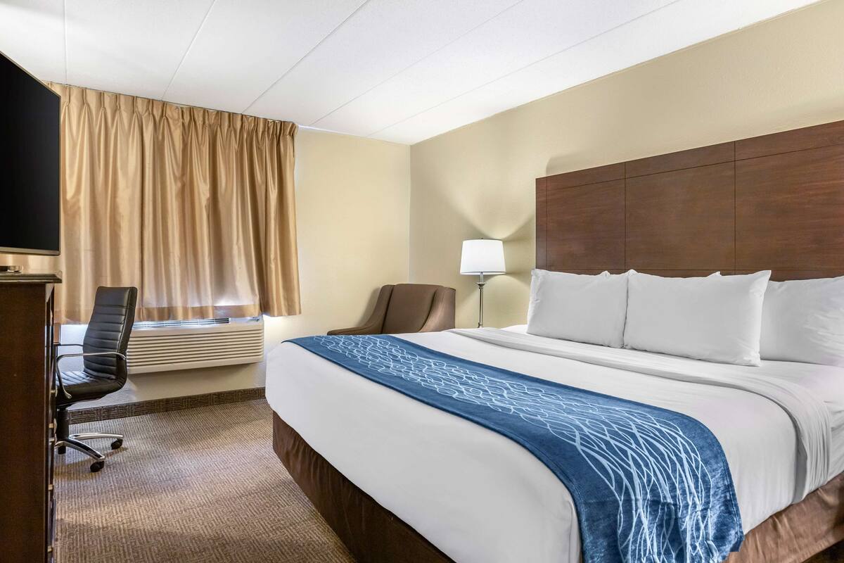 A hotel room with a large bed, a desk with a chair, a lamp, and a window with curtains.