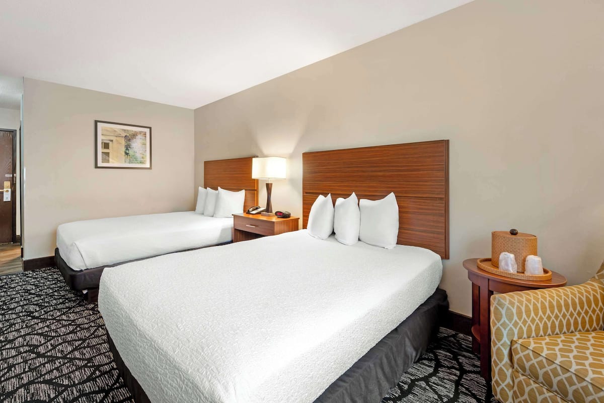 A hotel room with two beds, a bedside table with a lamp, a small side table with toiletries, and a patterned rug.