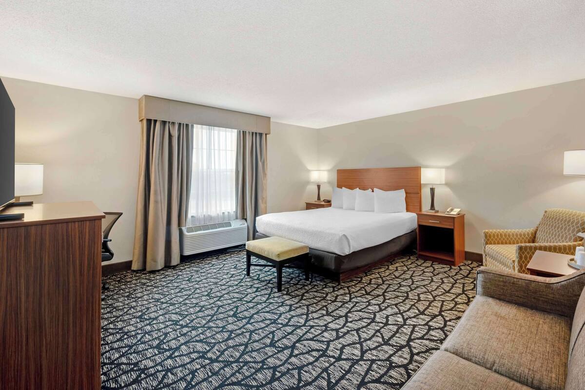 A hotel room with a bed, bedside tables, a desk, a chair, a small table, and a window with curtains.