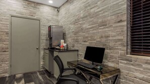 A modern office space with a glass desk, a black office chair, a computer, a refrigerator, and a window with blinds.