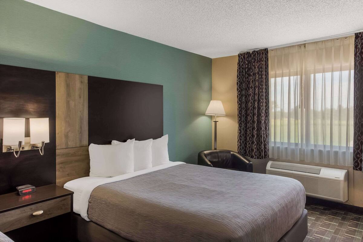 A hotel room with a large bed, a bedside table with a lamp, a chair, and an air conditioning unit.