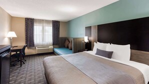 A hotel room with a large bed, a desk with a chair, a small seating area, and a window with curtains.