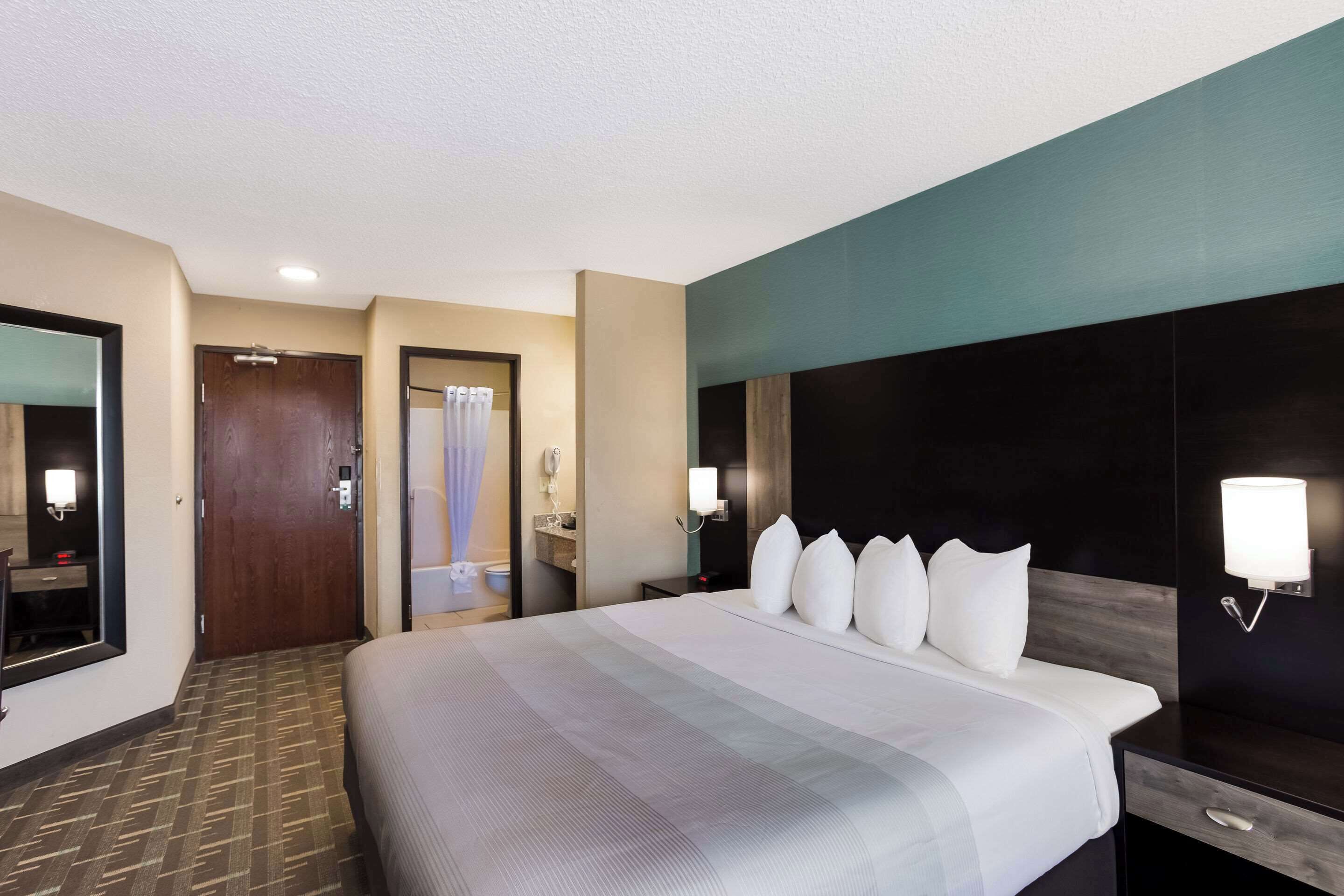 A hotel room with a large bed, a bedside table, a mirror, a wall-mounted light, and a door leading to the bathroom.