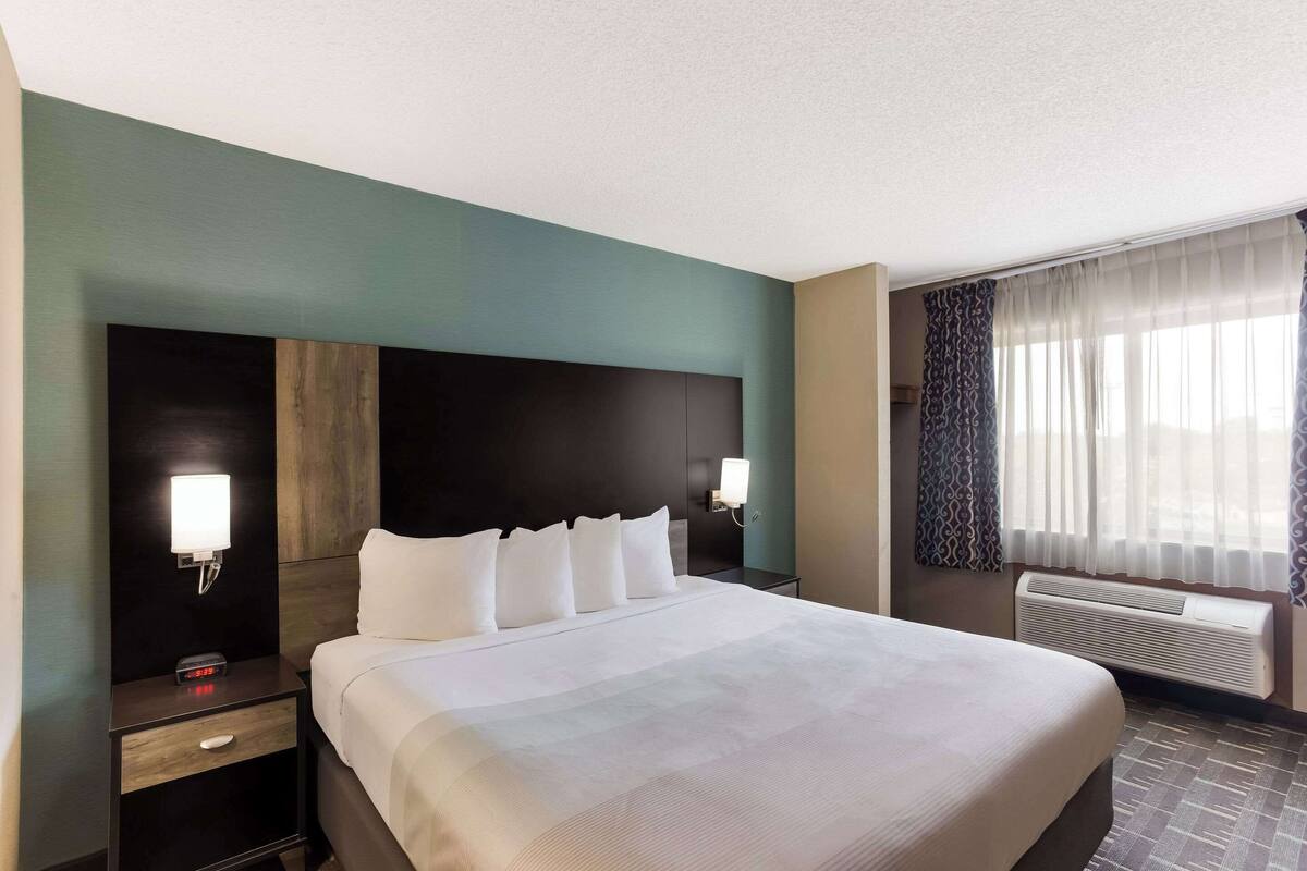 A hotel room with a large bed, a nightstand with a lamp, a wall-mounted heater, and a window with curtains.
