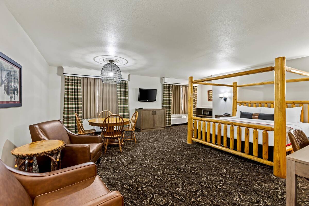 A hotel room with a four-poster bed, a dining area with a table and chairs, a television mounted on the wall, and a patterned carpet.