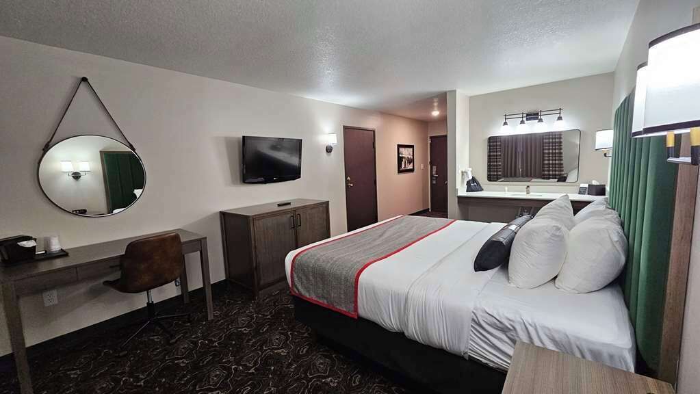Una habitación de hotel con una cama, un escritorio con silla, televisión, espejo y baño con lavamanos y espejo, además de una puerta.