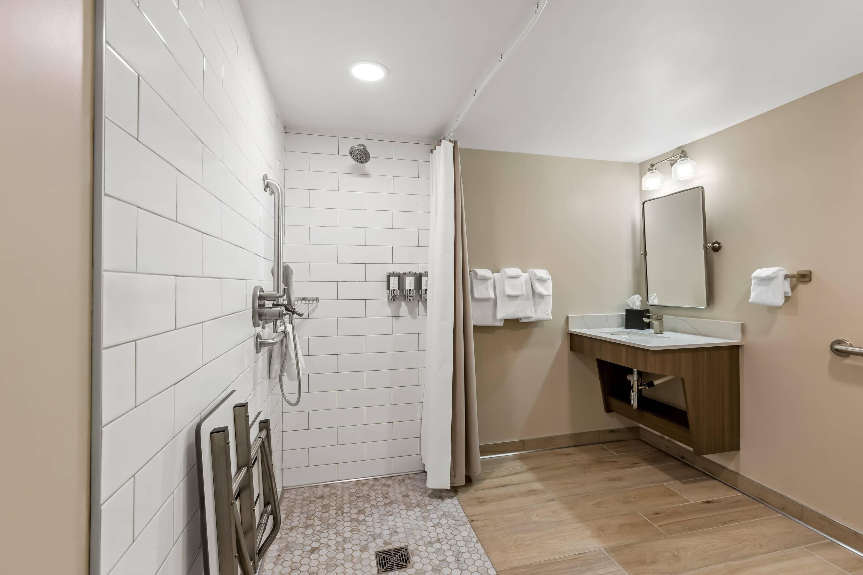 A modern bathroom with white tiled walls, a shower, a sink with a mirror, and a wooden vanity.