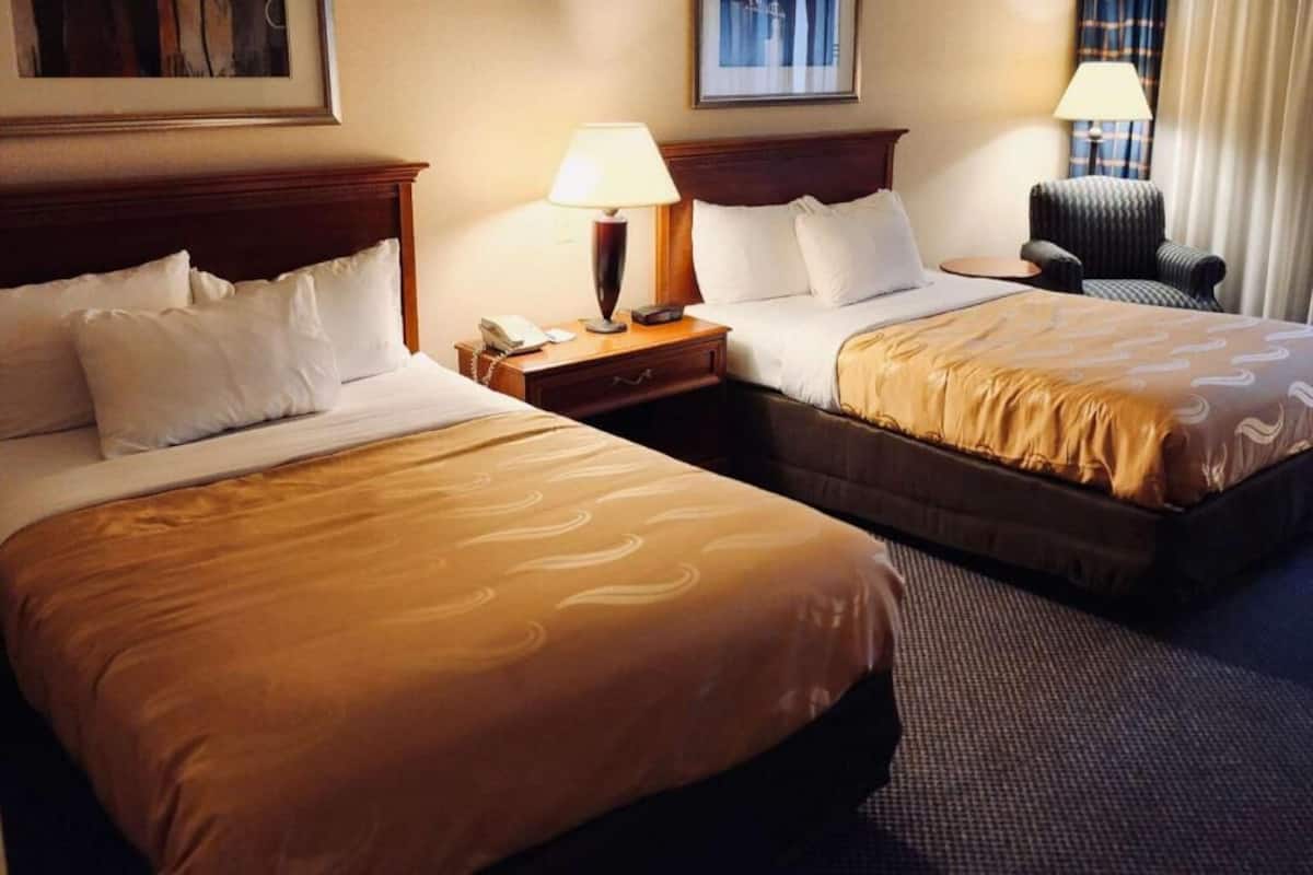 A hotel room with two beds, a nightstand with a lamp, a chair, and a framed picture on the wall.