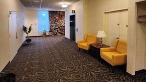 Een hotellobby met een kamerbreed tapijt, twee gele fauteuils, een lamp, een stenen muur en een lift.
