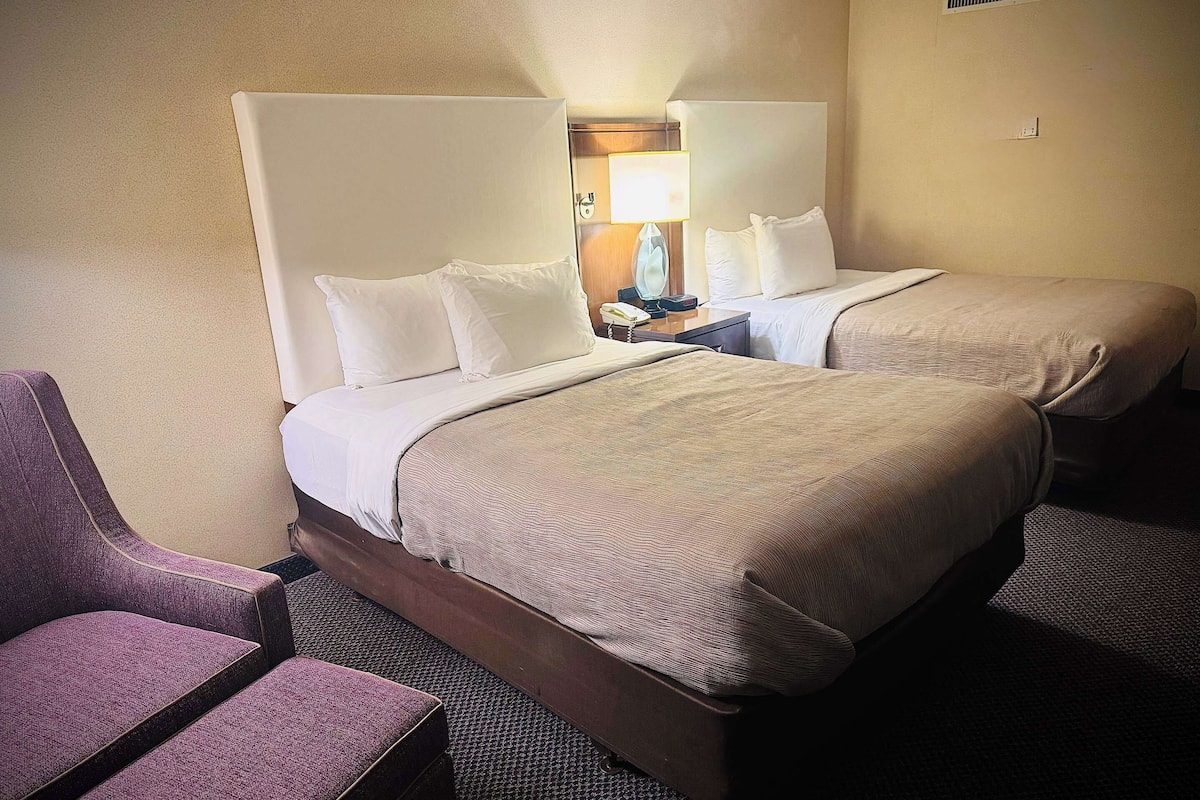 A hotel room with two beds, a chair, and a nightstand with a lamp.