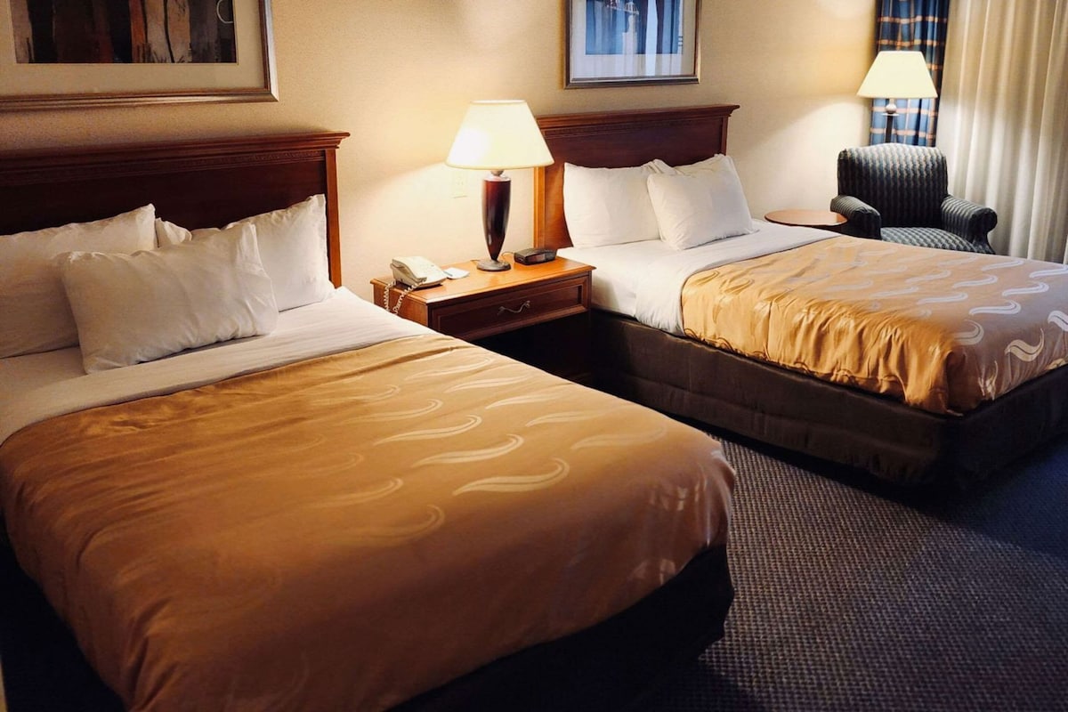 A hotel room with two beds, a nightstand with a lamp, a chair, and a framed picture on the wall.