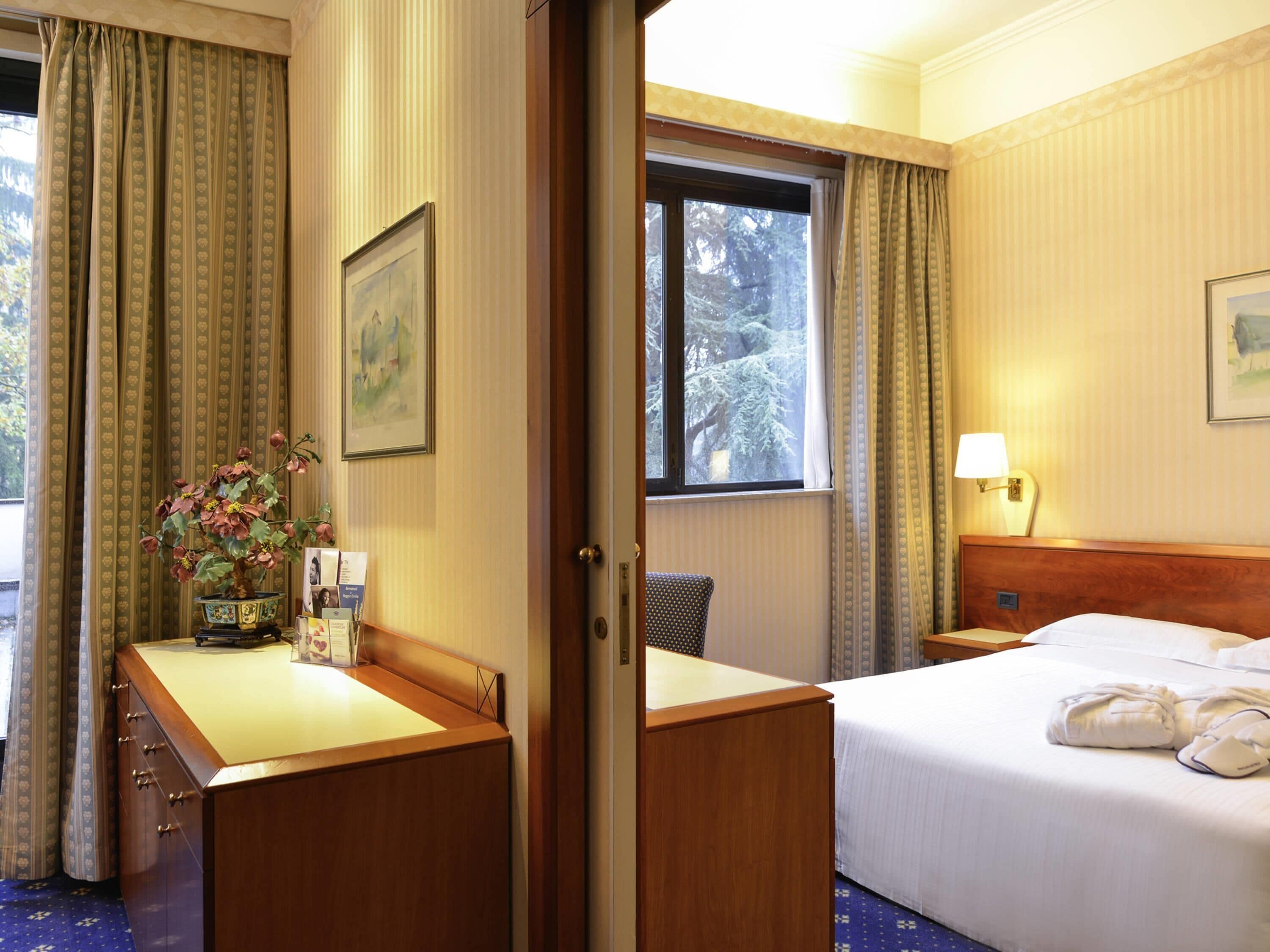 Habitación de hotel con cama, escritorio, silla, ventana y vista a árboles, además de una lámpara.