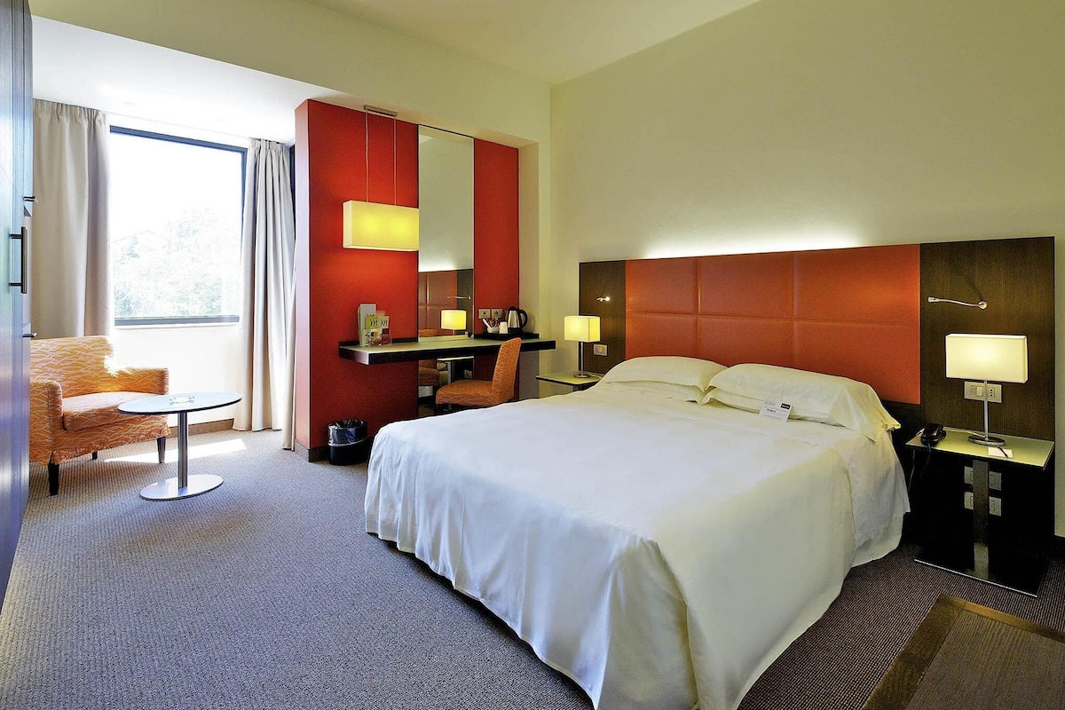 A hotel room with a bed, a desk, a chair, a desk lamp, a nightstand, a telephone, and a window with curtains.