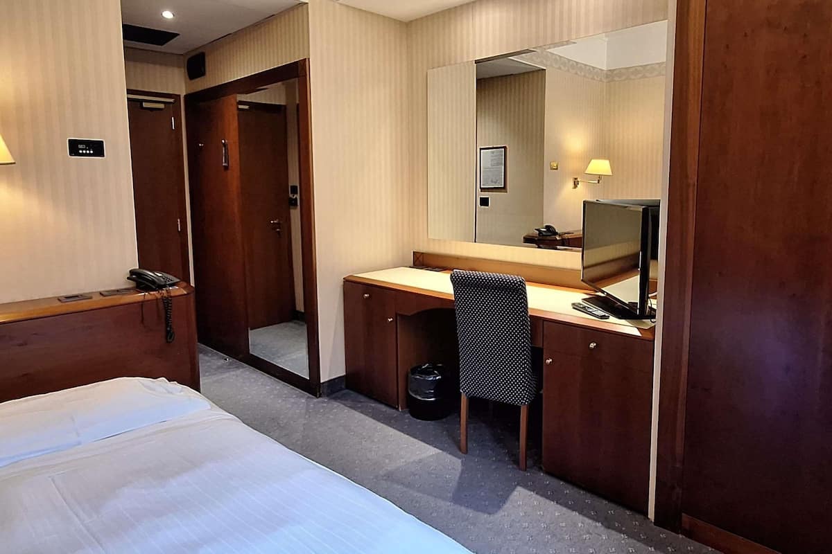 Ein Hotelzimmer mit einem Bett, einem Schreibtisch, einem Stuhl, einer Schreibtischlampe, einem Telefon, einem Papierkorb und einem Spiegel.