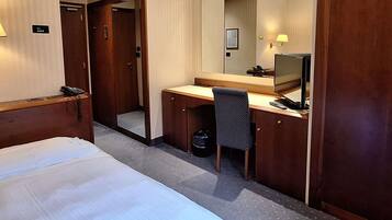 A hotel room with a bed, a desk, a chair, a desk lamp, a telephone, a trash bin, and a mirror.