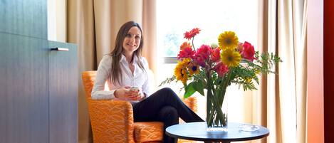 Una mujer sentada en una silla color naranja con estampado, en una habitación con una mesa redonda, un jarrón de flores y una ventana con cortinas.