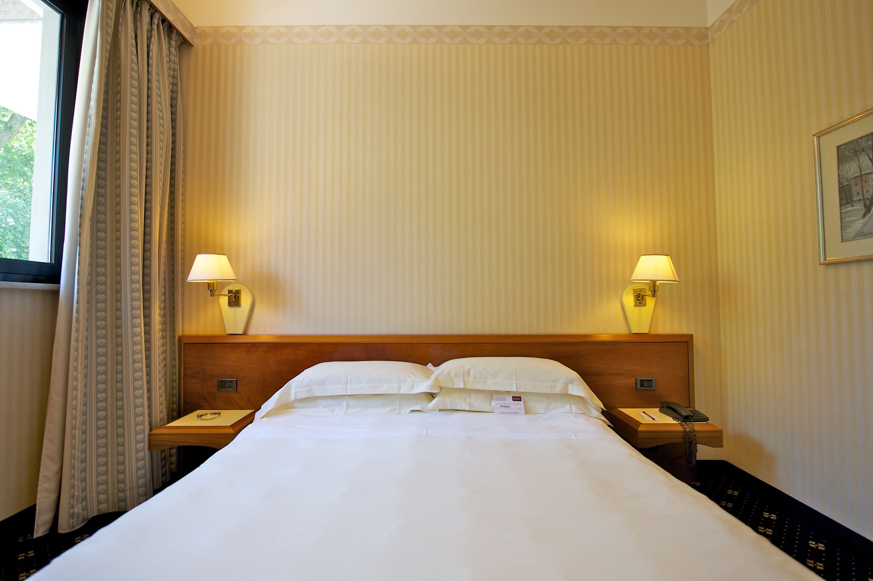 A hotel room with a large bed, two bedside tables with lamps, a wooden headboard, and a window with curtains.