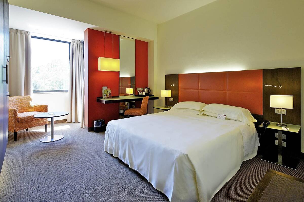 A hotel room with a bed, a desk, a chair, a desk lamp, a nightstand, a telephone, and a window with curtains.