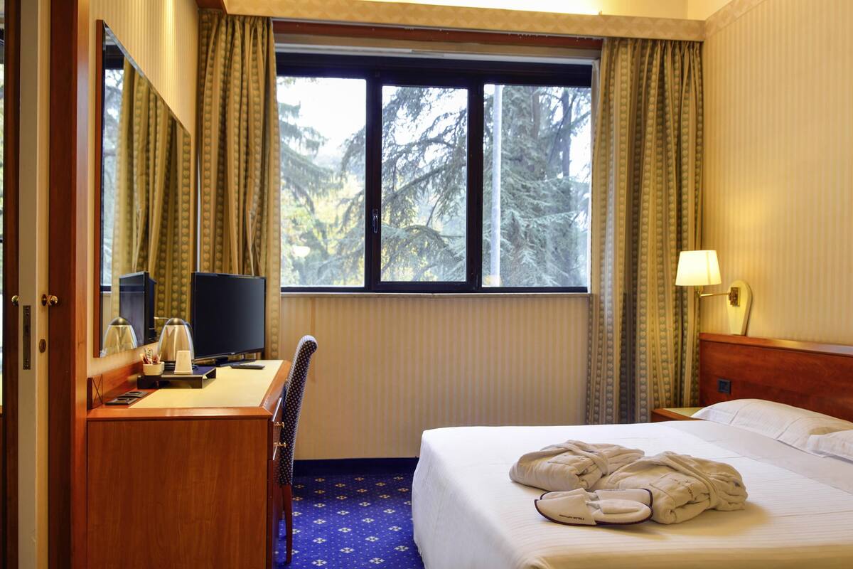 Ein Hotelzimmer mit Bett, Schreibtisch mit Computer, Stuhl, Fenster mit Vorhängen und einer Lampe.