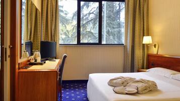 Camera d'albergo con un letto, una scrivania con un computer, una sedia, una finestra con tende e una lampada.