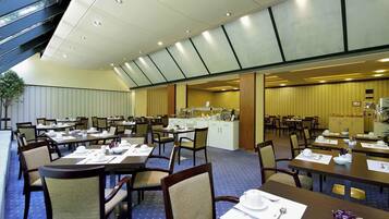 Ein großzügiger Hotel-Speisesaal mit mehreren Tischen und Stühlen, einer Buffet-Anrichtung und einem großen Fenster, das für natürliches Licht sorgt.