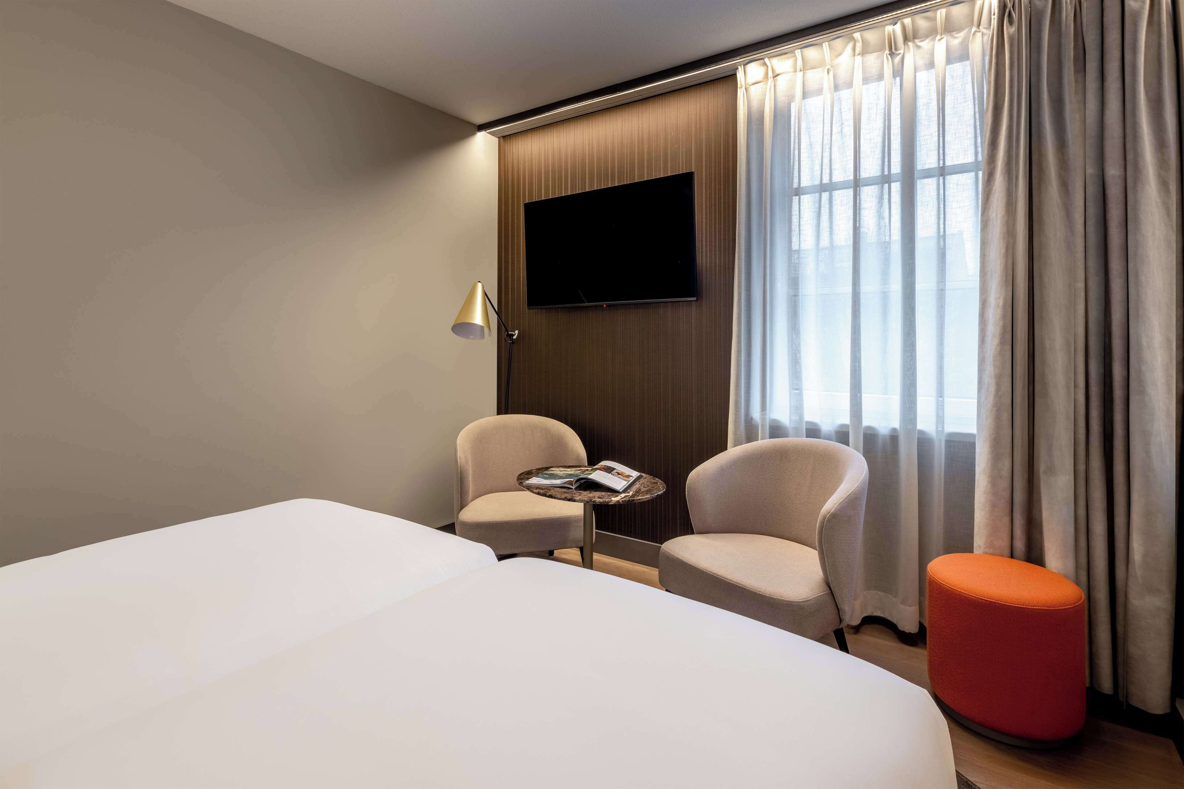 Ett hotellrum med en stor säng, två fåtöljer, ett litet bord med ett tidskriftsnummer, en tv som är monterad på väggen och ett fönster med gardiner.