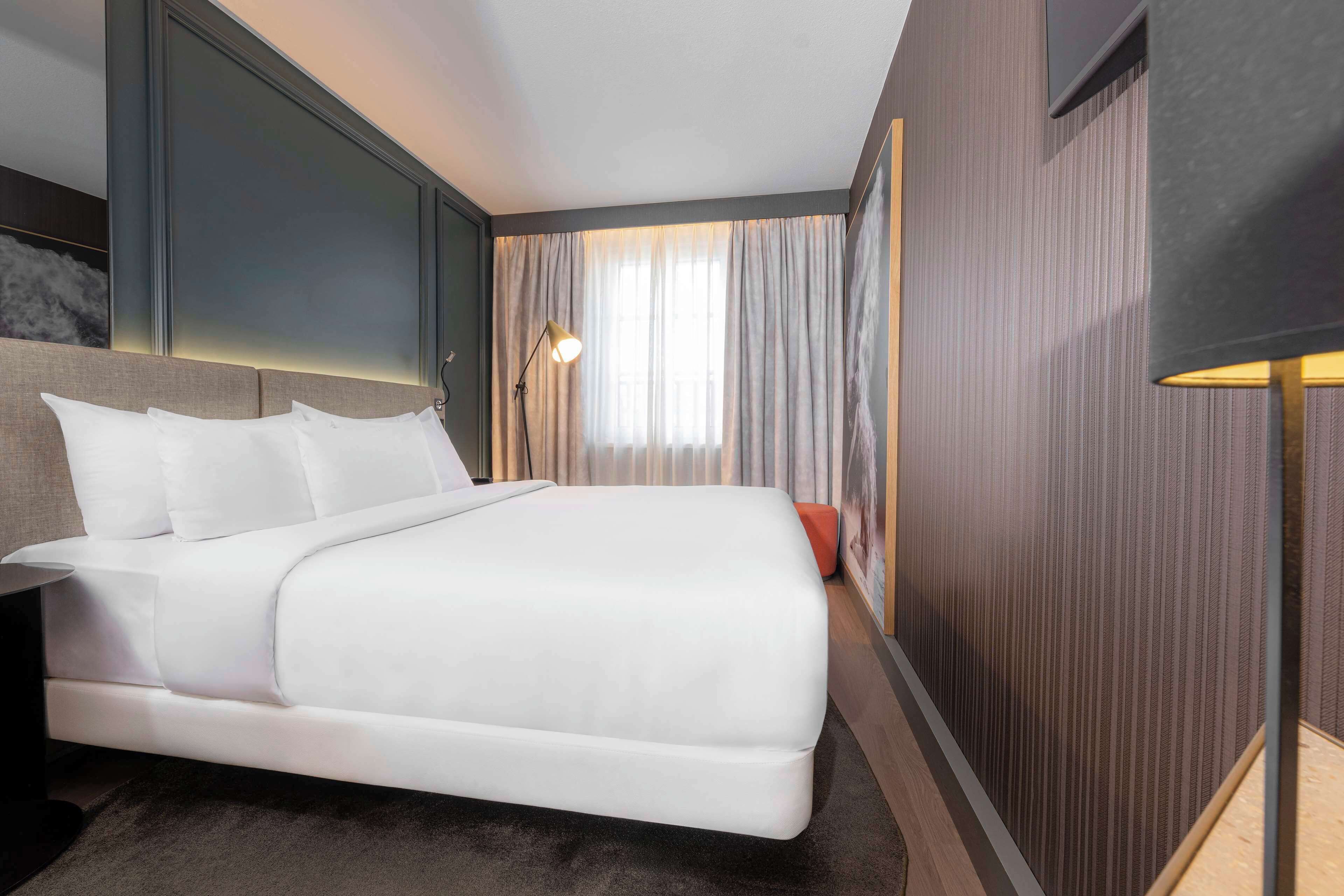 Ein modernes Hotelzimmer mit einem großen Bett, einem Nachttisch, einer Lampe und Blick ins Freie durch Vorhänge.
