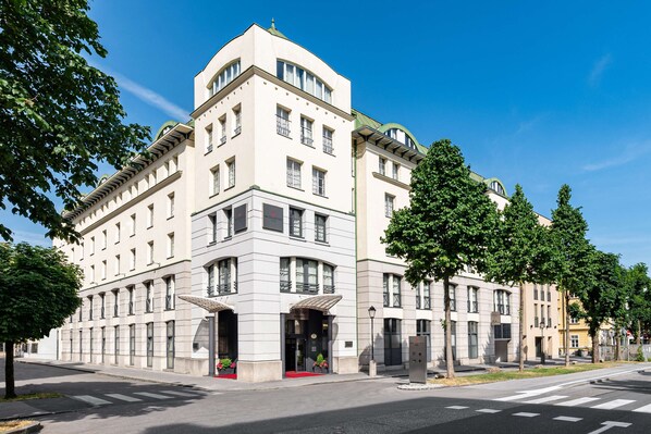Un grand hôtel à plusieurs étages, de style architectural classique, qui se distingue par son entrée d’angle imposante et un tapis rouge menant au hall d’entrée.