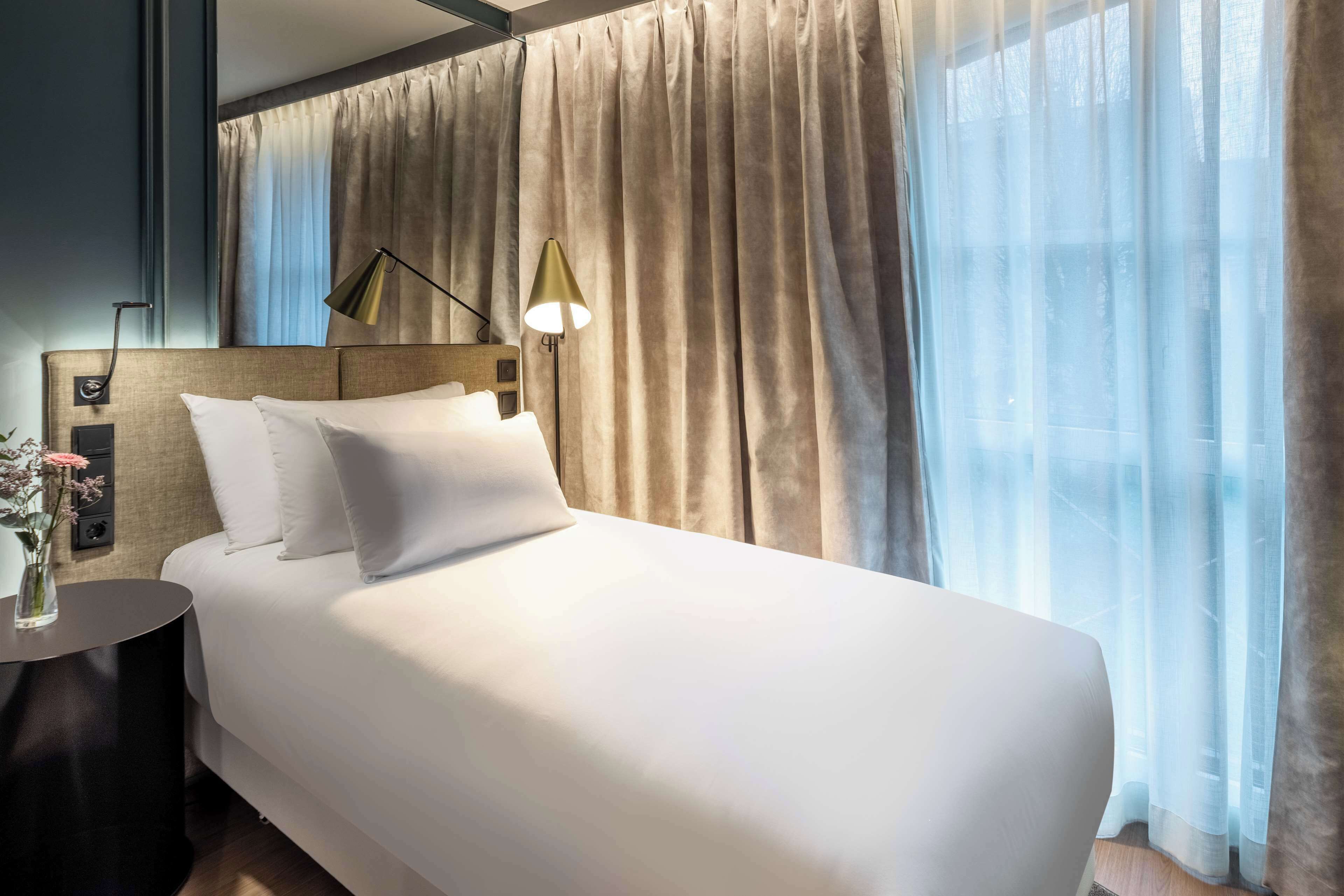 A hotel room with a large bed, a bedside table with a lamp, and a view of the outdoors through sheer curtains.