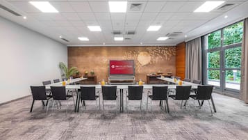 Ein Konferenzraum mit einem langen Tisch, Stühlen, einer Projektionsleinwand und einem großen Fenster mit Blick auf Grünflächen.
