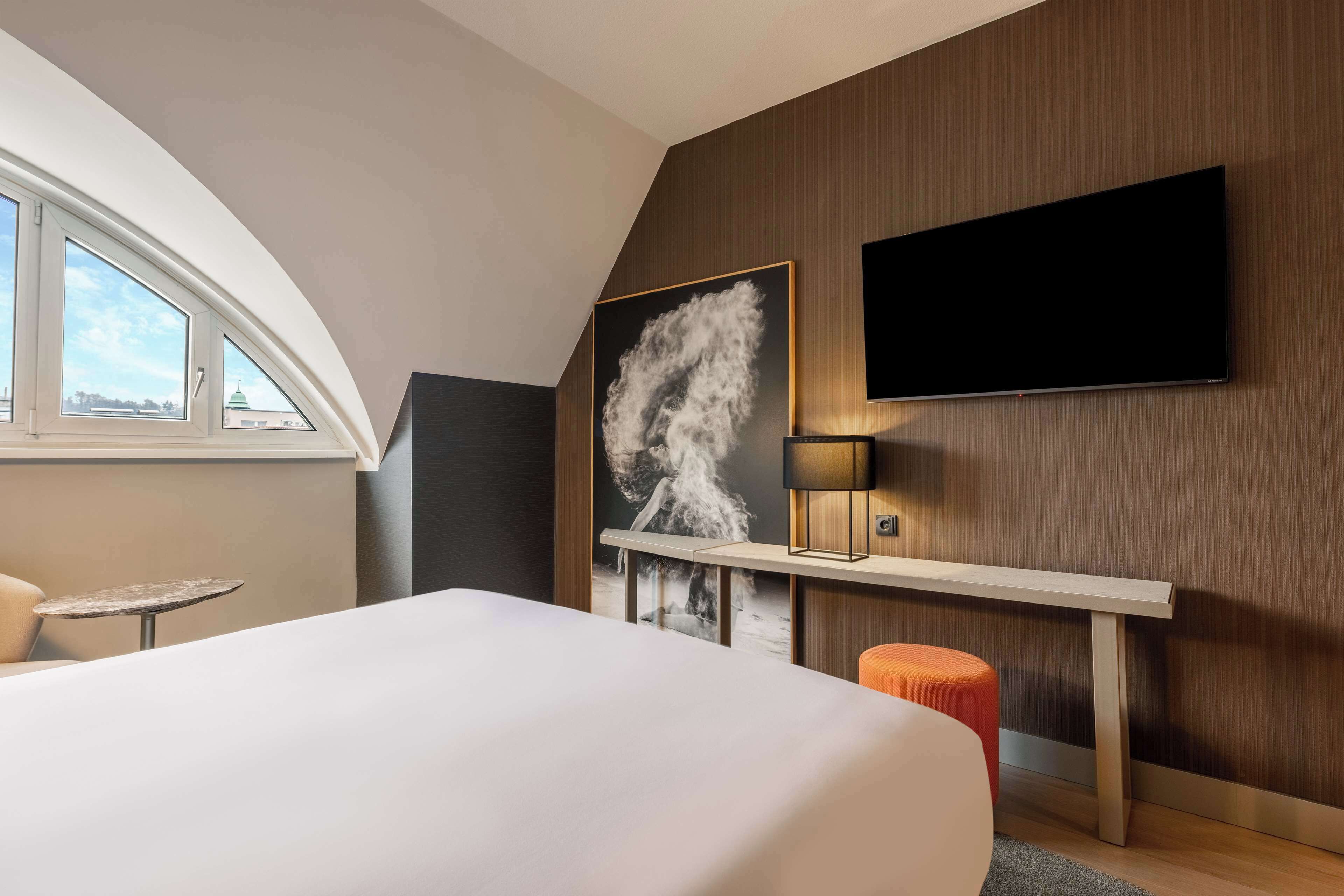 A modern hotel room with a large bed, a TV mounted on the wall, a painting, and a small table.