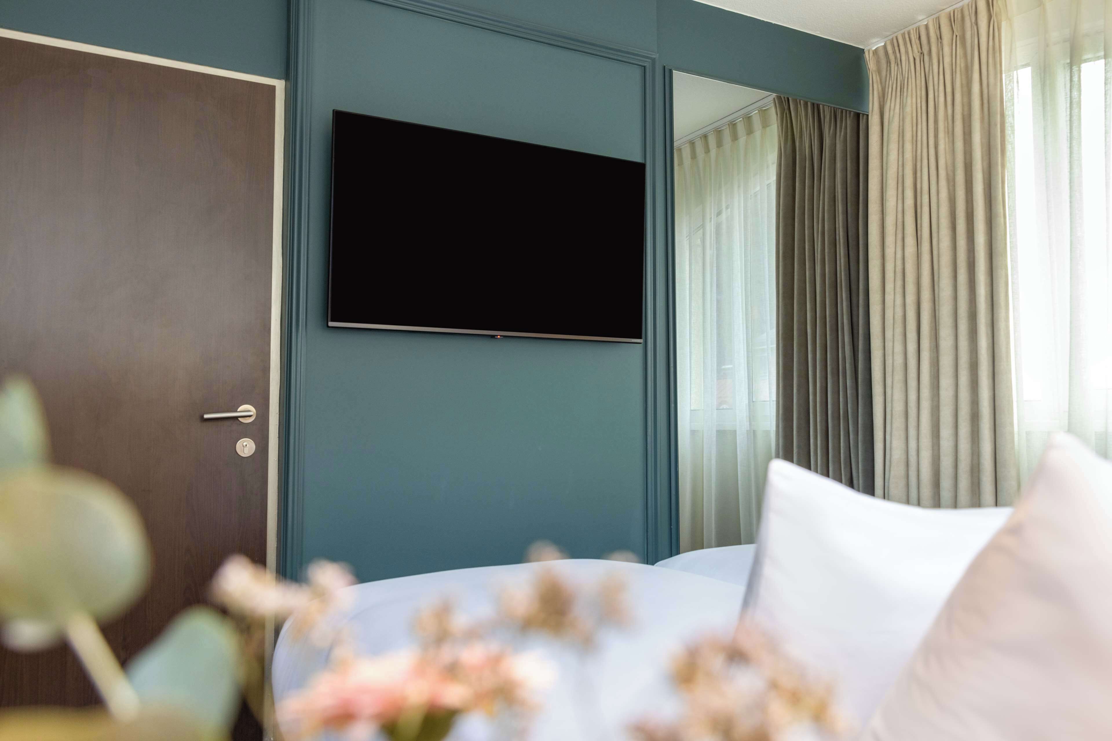A modern hotel room with a large flat-screen TV mounted on the wall, a bed with white bedding, and a window with sheer curtains.