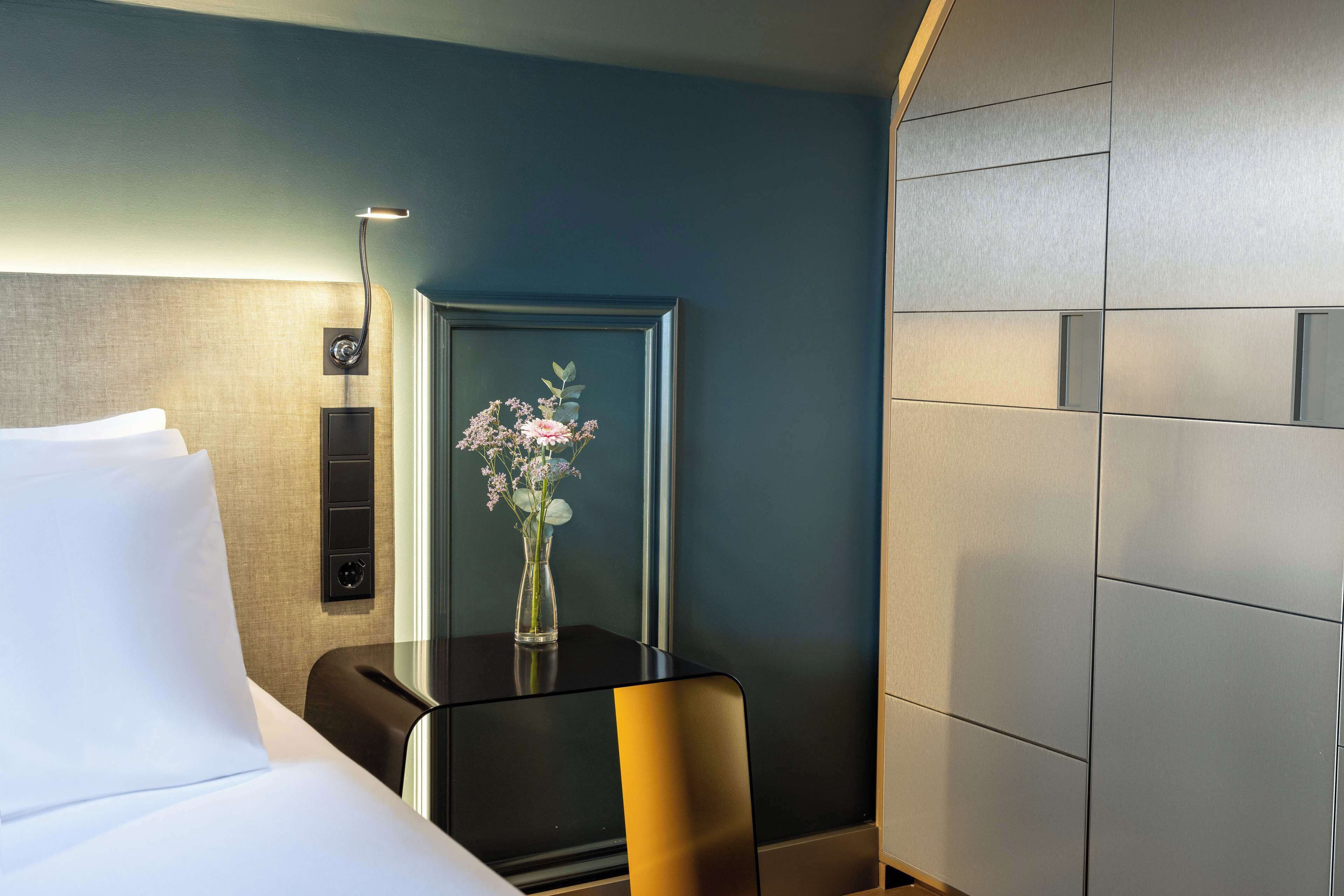 A hotel room with a bed, a bedside table with a vase of flowers, a wall-mounted clock, and a modern light fixture.