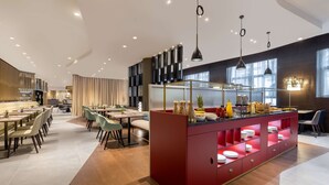 A modern dining area with multiple tables and chairs, a central island with a red base, and a kitchen area with a glass partition.